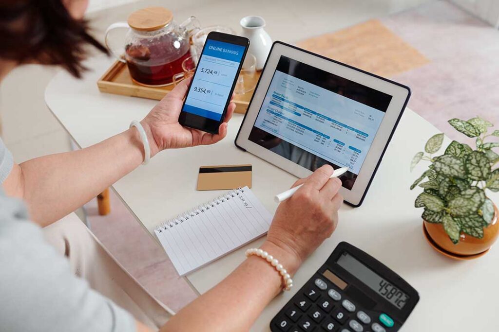 A person reviewing their bank account on a smartphone and checking a printed account statement at a home table.